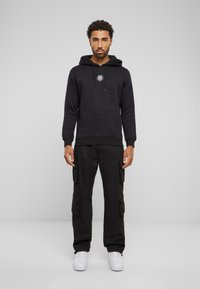 Zwarte hoodie met een buidelzak en een capuchon met trekkoord, voorzien van een rond logo. Gecombineerd met zwarte cargo pants en witte sneakers.
