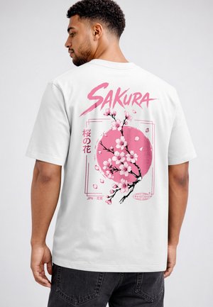 Man wearing white t-shirt with pink cherry blossom branch design and text "SAKURA" on back against plain background.