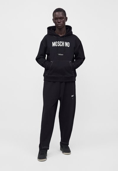 Άνδρας φοράει μαύρο φούτερ Moschino με λευκό λογότυπο και ταιριαστό μαύρο παντελόνι και αθλητικά παπούτσια σε απλό φωτεινό φόντο.
