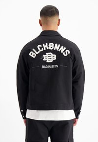 Zwarte denimjas met geborduurde tekst op de rug: "BLCKBNNS" en "BAD HABITS" in contrasterende witte draad en een cropped fit.