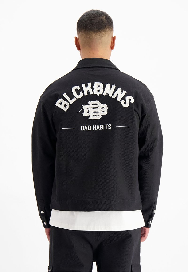 Zwarte denimjas met geborduurde tekst op de rug: "BLCKBNNS" en "BAD HABITS" in contrasterende witte draad en een cropped fit.