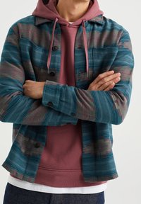 Teal- och flerfärgat mönstrat skjorta med knappar över en rosa hoodie, med synlig strukturerad textil och kontrasterande färger. Avslappnad lager-utseende.