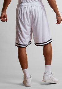 Shorts de sport blancs avec des rayures noires à l'ourlet, une taille élastique et un tissu léger. Portés avec des baskets blanches et des chaussettes.