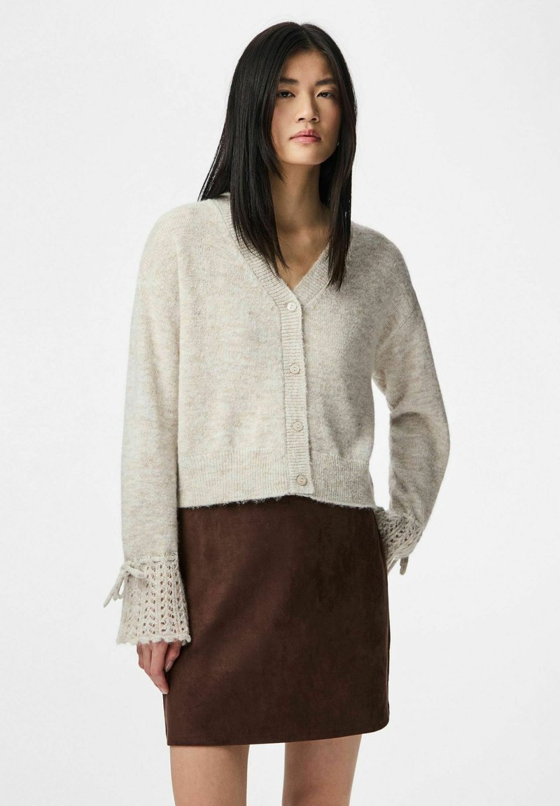 Woman wearing beige buttoned cardigan with lace cuffs and brown suede skirt, standing against a plain white background.