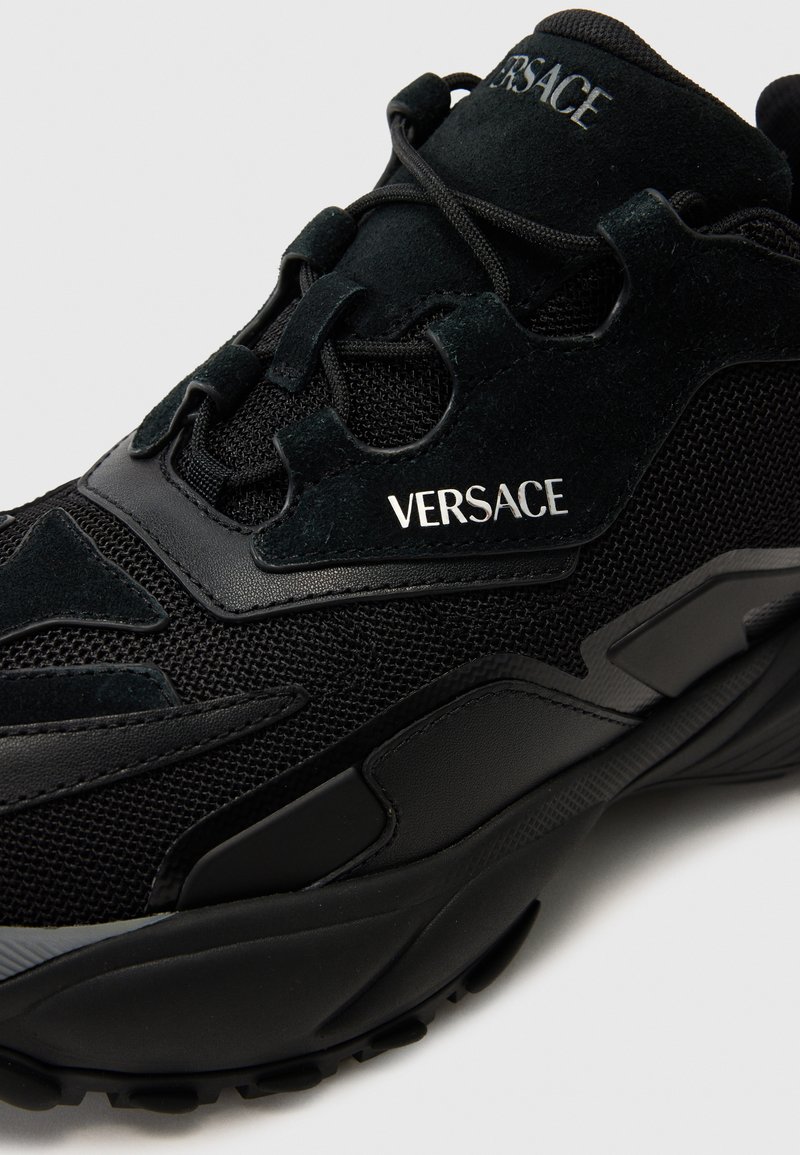 Zwarte sneaker met een combinatie van mesh en suède materialen, een chunky silhouet en een glanzend "VERSACE" logo aan de zijkant. Geprofileerde zool voor grip.