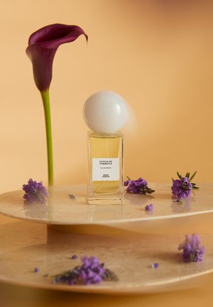 Klares Glas-Parfümfläschchen mit weißem rundem Verschluss, gefüllt mit gelber Flüssigkeit. Platziert auf einer Marmoroberfläche mit violetten Blumen und einer Calla-Lilie.