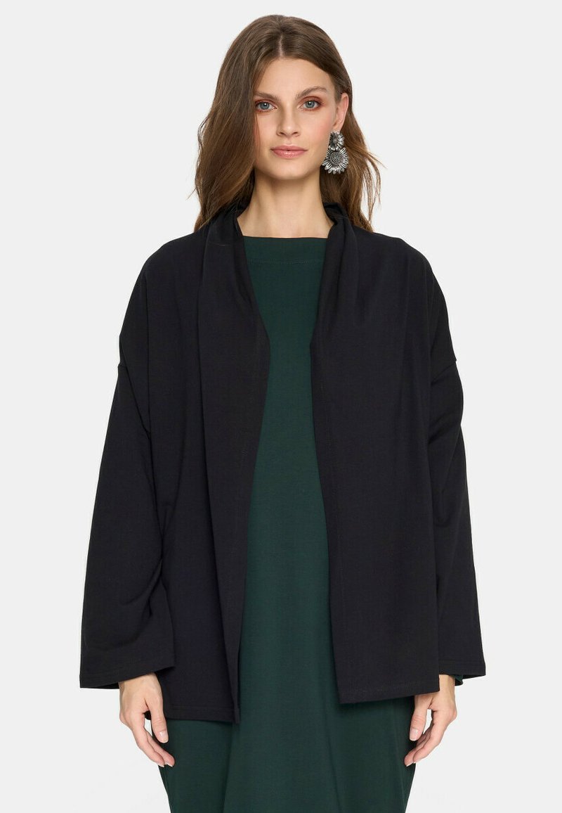 Czarna, oversize kurtka z kołnierzem typu szal, szerokimi rękawami i luźnym krojem, uszyta z miękkiego materiału, noszona na ciemnozielonej sukience.