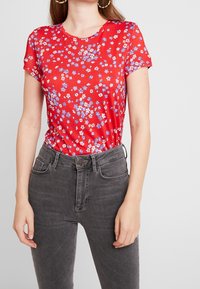 Red short-sleeve t-shirt with a floral pattern in blue and white, paired with high-waisted dark gray jeans. Smooth fabric, fitted design.