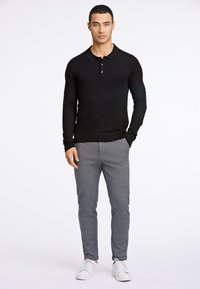 Svart langermet polo-skjorte med ribbestrikket nederkant, kombinert med grå teksturerte bukser og hvite joggesko. Enkelt design, nøytrale toner.