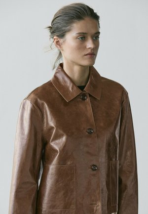 Kvinde med samlet hår, iført en brun læderjakke med knapper og store lommer, som tænksomt kigger til siden mod en ensfarvet baggrund.