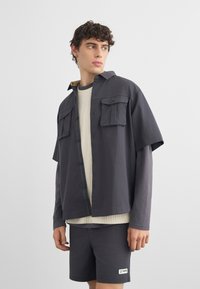 Νέος άνδρας με σγουρά μαλλιά που φορά στρωμένα γκρι και κρεμ καθημερινά ρούχα, κοιτάζει στο πλάι έναν απλό φόντο.