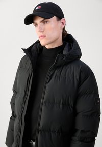 Black puffer jacket with a hood, featuring a zip front and insulated sections. Paired with a black corduroy cap with a logo accent.