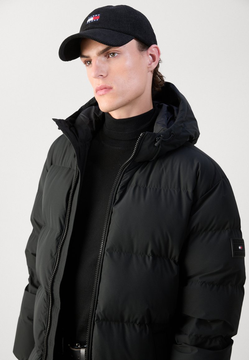 Chaqueta de plumas negra con capucha, con cierre de cremallera en la parte delantera y secciones aislantes. Combinada con una gorra de pana negra con un acento de logo.