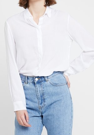 Femme portant une chemise blanche à manches longues rentrée dans un jean bleu clair taille haute, une main sur la hanche.