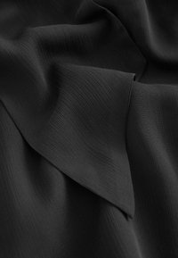 Čierna tkanina s textúrovaným, rebrovaným povrchom, so hladkými, voľne visiacimi závesmi a prehnutým okrajom. Celkový vzhľad je mäkký a ľahký.