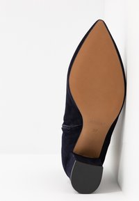Chaussure en daim noire à bout pointu vue de la semelle, présentant une semelle lisse couleur tan et un talon bloc, taille 37 indiquée sur la semelle.