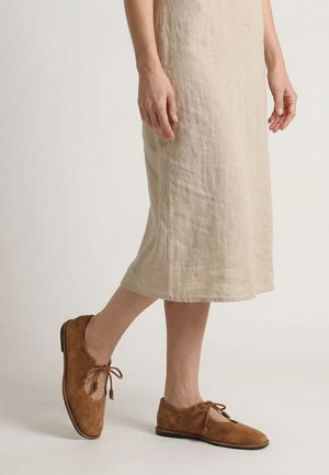 Vrouw draagt een beige midi rok en bruine suède veterschoenen met een platte zool, staand op één been tegen een lichte achtergrond.