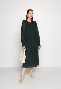 Vestido longo de mangas compridas e pregas, na cor verde escuro, com detalhes em babados. Combinado com uma bolsa bege clara e sapatos de salto aberto com tiras.