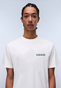Hvid bomulds t-shirt med et blåt og sort trykt logo på venstre bryst. Enkel rund halsudskæring og korte ærmer.