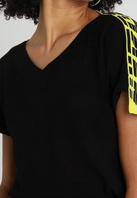 Top noir à col V avec une texture en maille, présentant une accentuation rayée jaune vif sur l'épaule. Manches courtes.