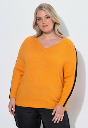 Donna con capelli biondi che indossa un maglione arancione a V con strisce nere sulle maniche e pantaloni neri, in piedi davanti a uno sfondo chiaro.