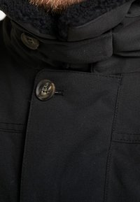 Detailní záběr na límec černého zimního kabátu s knoflíky a fleece podšívkou, přičemž částečně viditelný je vous.
