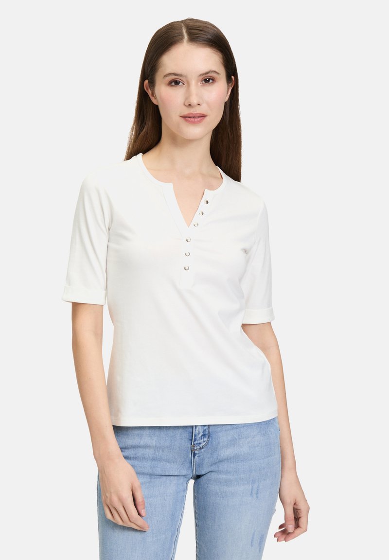 Chemise blanche en coton à manches courtes avec un col mandarin, comportant cinq boutons décoratifs et des manches relevées, associée à un jean bleu clair.