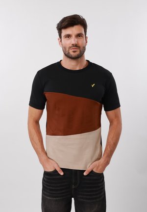 Άνδρας με σκούρα μαλλιά και γένια που φοράει μπλουζάκι με μπλοκ χρωμάτων σε μαύρο, καφέ και μπεζ, στέκεται με τα χέρια στις τσέπες μπροστά σε ένα απλό φόντο.