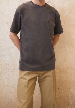 Mężczyzna ubrany w jednolity ciemnoszary t-shirt z krótkim rękawem i małym logo na piersi, w połączeniu z beżowymi spodniami o prostych nogawkach, stoi na tle teksturowanej ściany.