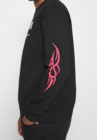 Sudadera negra con un gran diseño tribal rosa en la manga derecha, puños acanalados y cuello redondo.