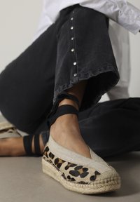 Leopardprint espadrille met een beige canvas bovenkant, geweven jute zool en zwarte enkelbandjes, geplaatst tegen donkere broek met metallic knopen.