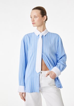 Donna con capelli raccolti in uno chignon, indossa una camicia a righe azzurre e bianche con bottoni, cravatta bianca, pantaloni bianchi e orecchini d'oro, guarda di lato.
