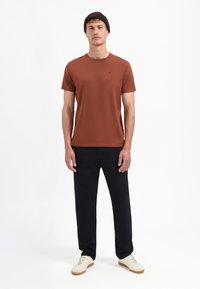 Roodbruine T-shirt met korte mouwen en een ronde hals, gecombineerd met zwarte broek en witte sneakers. Eenvoudig ontwerp, minimale merkdetails.