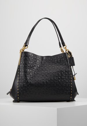 Sac à main en cuir noir avec motif embossé, clous dorés sur les côtés, double poignée et petite étiquette noire attachée.