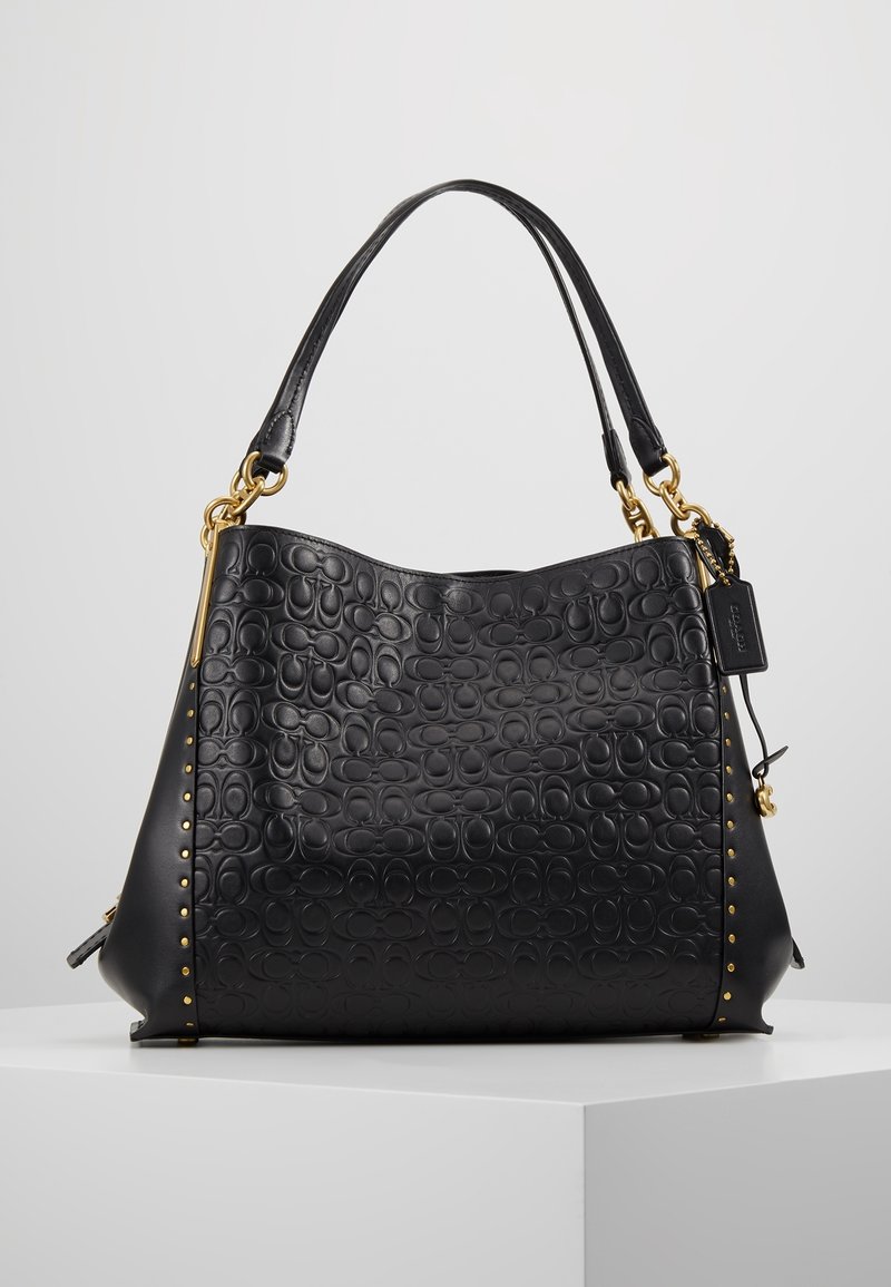 Sac à main en cuir noir avec motif embossé, clous dorés sur les côtés, double poignée et petite étiquette noire attachée.
