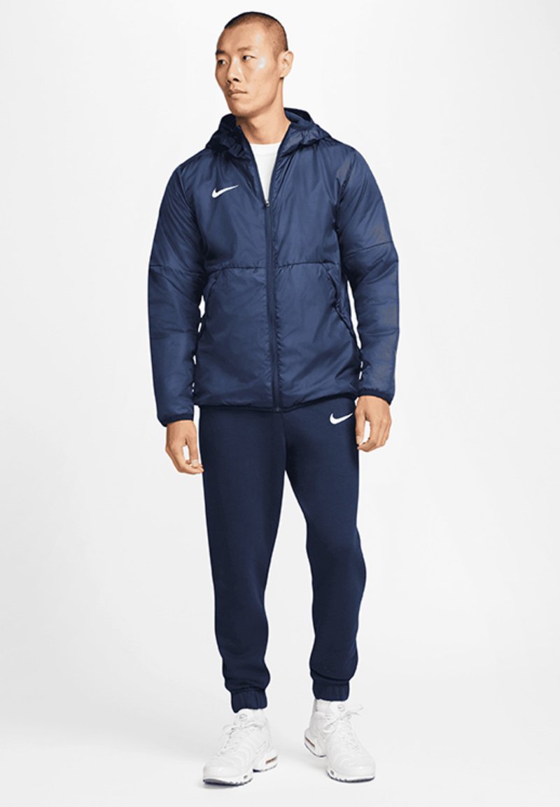Marineblauwe geïsoleerde jas met een capuchon, rits aan de voorkant en elastische manchetten, gecombineerd met marineblauwe trainingsbroek; gedragen met witte sportschoenen.