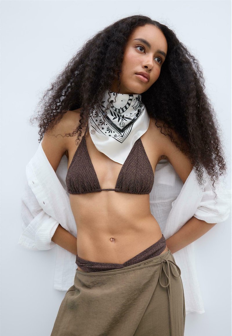 Femme aux longs cheveux bouclés, portant un haut de bikini marron, un foulard blanc à motif autour du cou, une chemise blanche ouverte et une jupe portefeuille olive, posant les mains dans le dos.