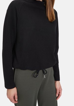 Žena nosící černý top s dlouhým rukávem a olivově zelené kalhoty se šňůrkou, stojí s jednou rukou uvolněně podél těla.