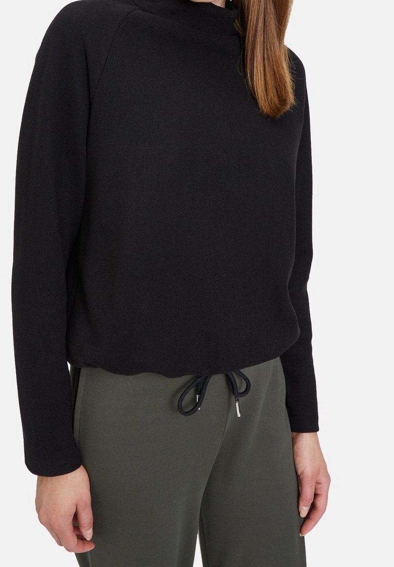 Femme portant un haut noir à manches longues et un pantalon vert olive à cordon, debout avec une main relâchée le long du corps.