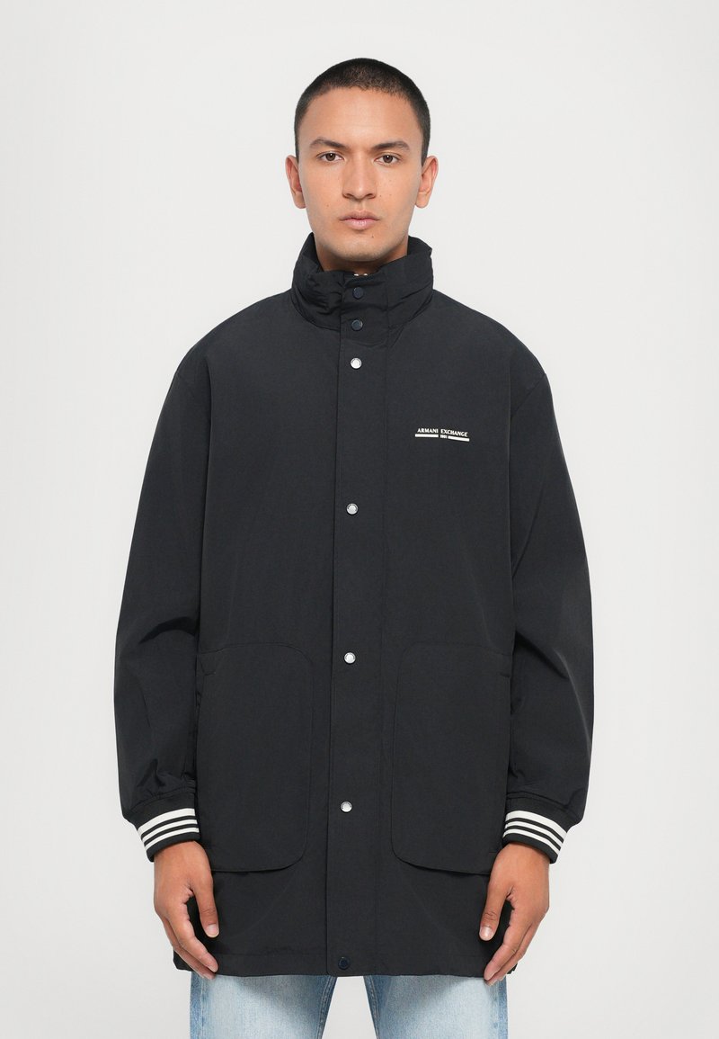 Black waterproof jacket with a high collar, snap buttons, two front pockets, and striped cuffs. Features a logo on the chest.