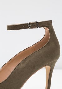 Olivově zelená semišová lodička na vysokém podpatku s úzkým páskem kolem kotníku, stříbrnou sponou a hladkým vnitřním potahem, s špičatou špičkou a elegantním tvarem.