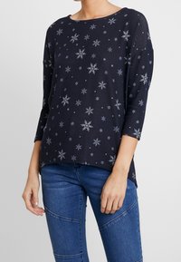 Haut à manches longues bleu marine avec motifs de flocons de neige blancs, encolure ronde, coupe décontractée et tissu doux. Associé à un jean en denim.