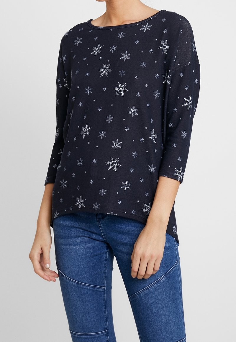 Haut à manches longues bleu marine avec motifs de flocons de neige blancs, encolure ronde, coupe décontractée et tissu doux. Associé à un jean en denim.
