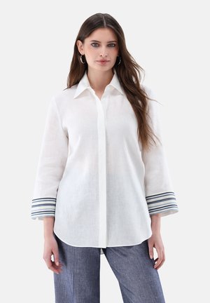 Junge Frau mit langen braunen Haaren, trägt Creolen, ein weißes Hemd mit gestreiften Manschetten und blaue Hose, steht vor weißem Hintergrund.