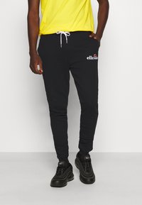 Sorte joggingbukser med elastisk talje og snøre, prydet med et rødt og hvidt logo på låret, parret med sorte sneakers og en gul skjorte.