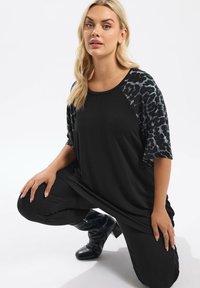 Schwarzes T-Shirt mit kurzen Ärmeln im schwarzen Leopardenmuster, aus weichem Stoff, lässige Passform. Getragen mit schwarzen Hosen und schwarzen High Heels.