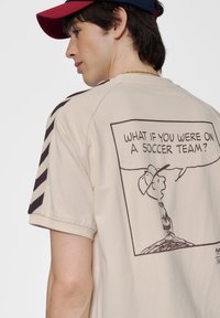 Crème kleurige t-shirt met korte mouwen, voorzien van een cartoonafbeelding op de achterkant. Het ontwerp bevat een zwarte omtrek en tekst in een spraakwolk.