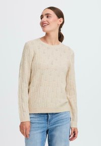 Lys beige strikket genser med et teksturert mønster og åpen vev, rund hals og ribbede mansjetter, sammen med blå jeans.