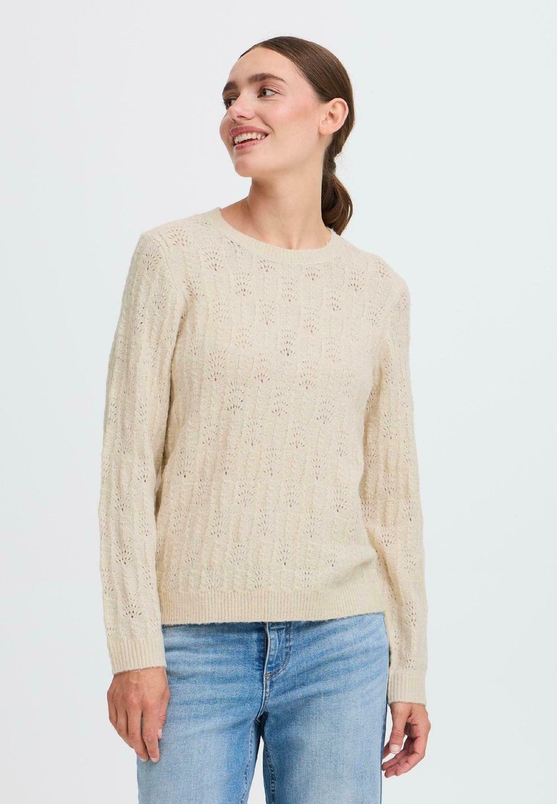 Lys beige strikket genser med et teksturert mønster og åpen vev, rund hals og ribbede mansjetter, sammen med blå jeans.