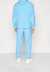 Lichtblauwe sporttracksuit van soepel materiaal, met een hoodie-jack en elastische manchetten, gecombineerd met bijpassende taps toelopende broek en witte sneakers.